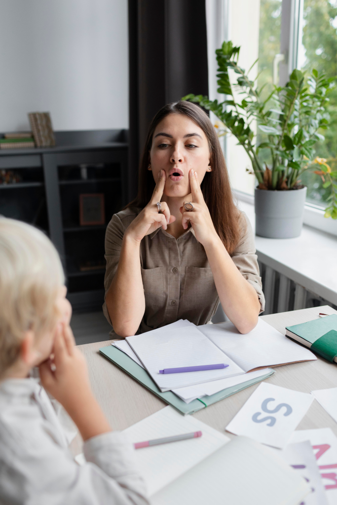 Speech therapist demonstrating mouth movements to a child during a therapy session at a table with notebooks and a plant nearby.