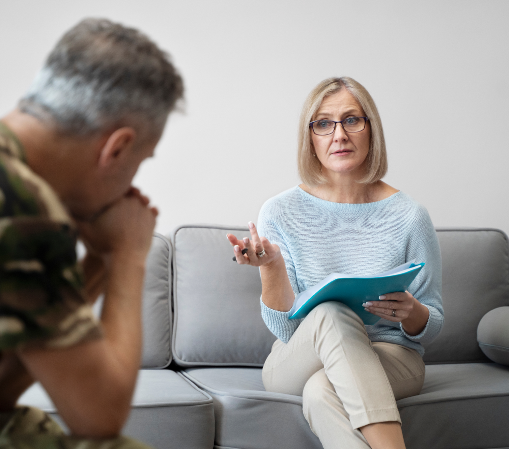 Woman with glasses holding a blue folder talking to a man in military uniform seated on a couch.