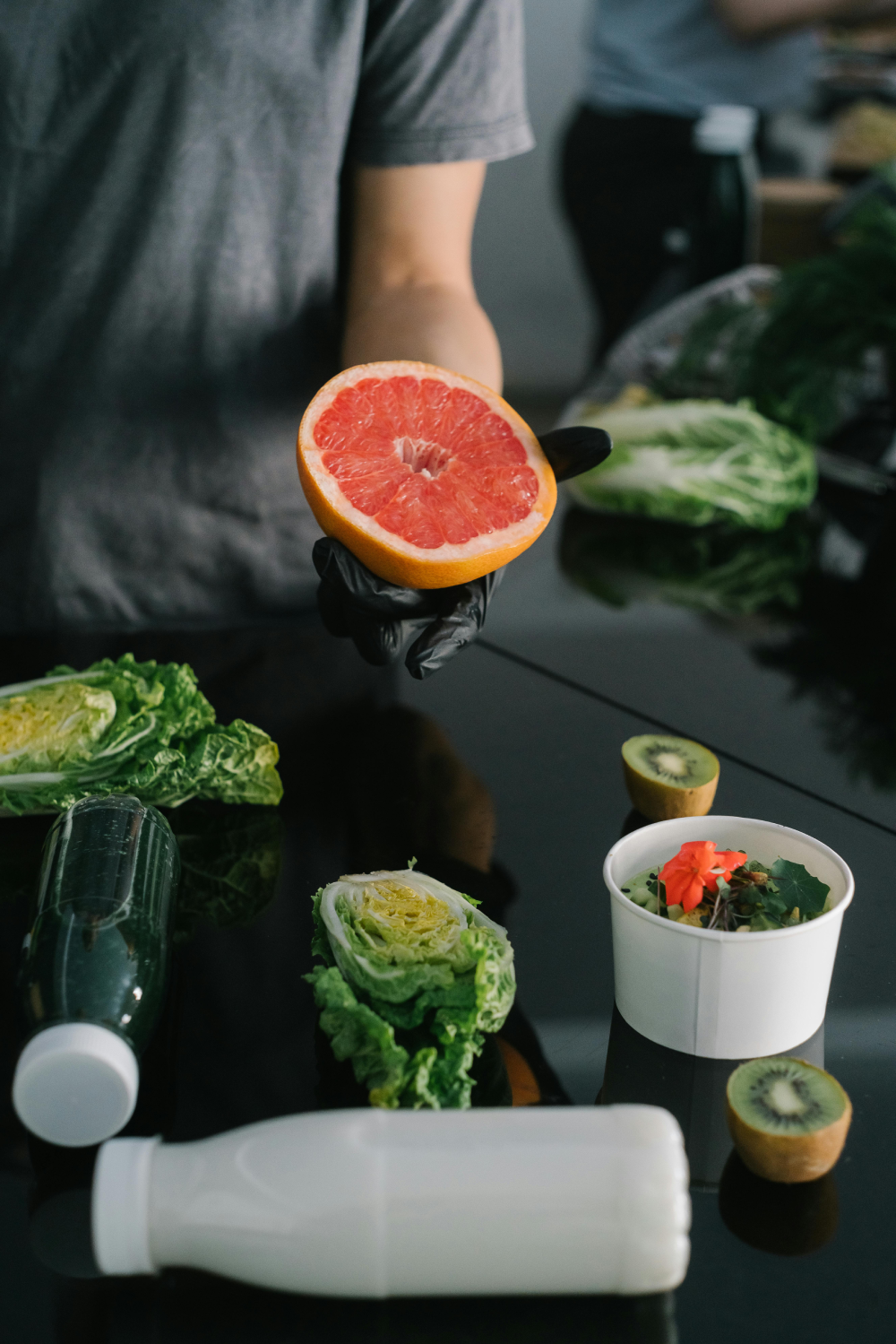 Person wearing a gray shirt and black glove holding half a grapefruit above a table with leafy greens, kiwis, a white bottle, and a green drink bottle.