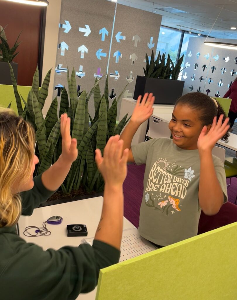 A smiling young girl and a woman facing each other with raised hands, about to high-five in an indoor office setting with plants and glass partitions.
