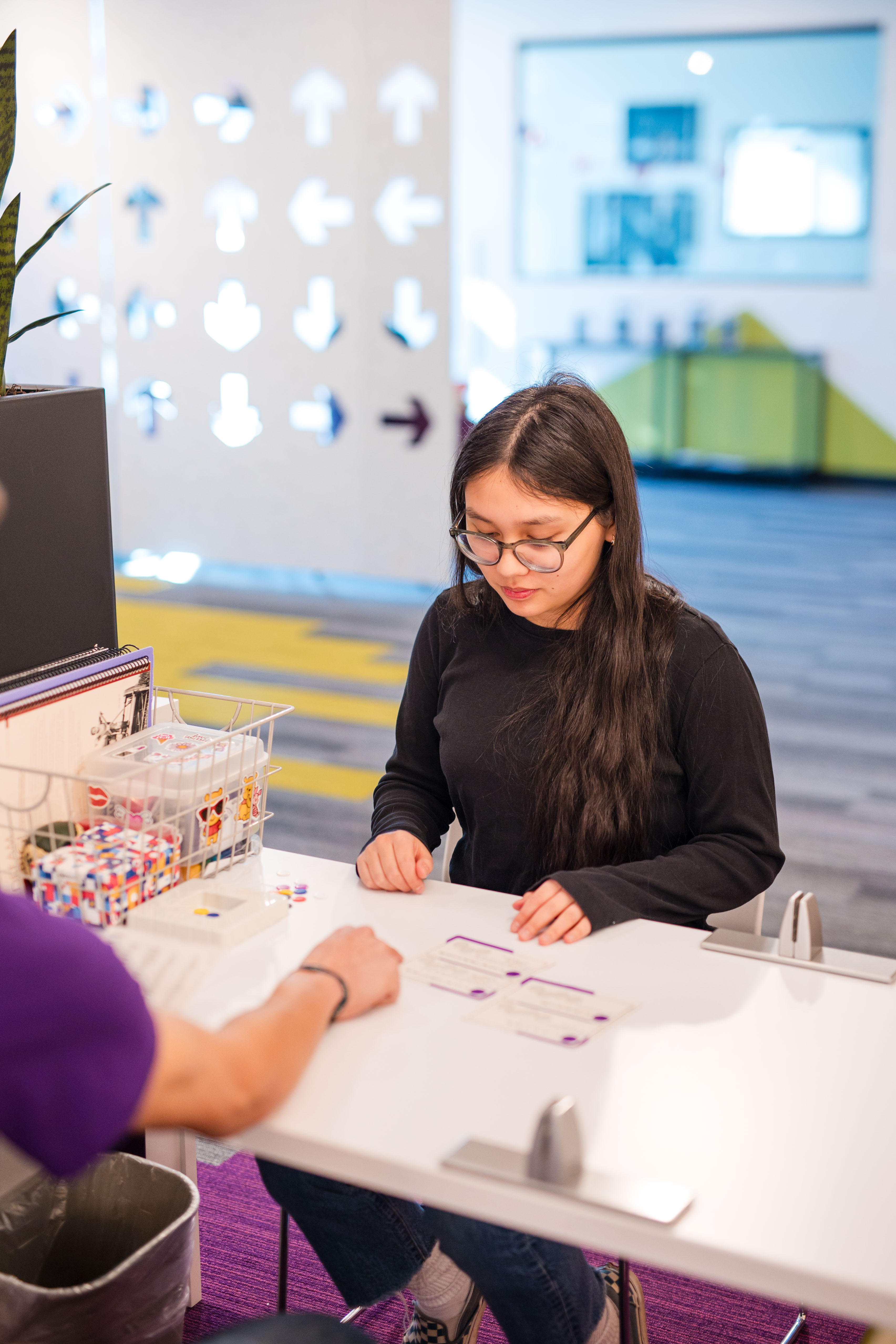Young woman with glasses sitting at a white table, engaging in a discussion with another person whose hand is visible, in a brightly lit modern office space.