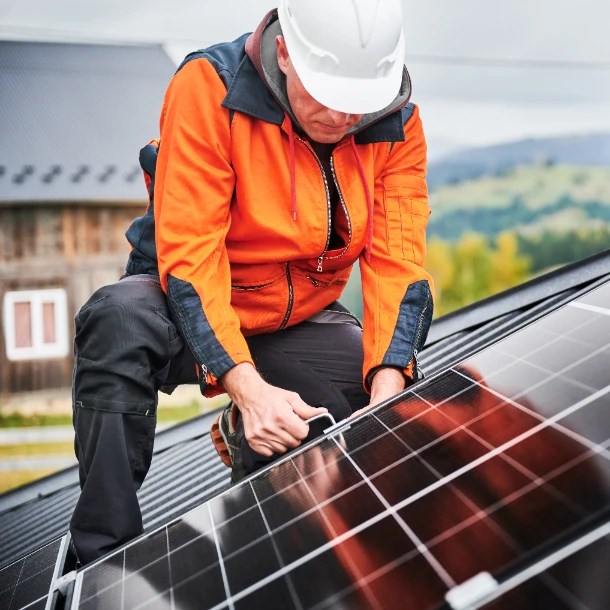 Técnico con casco blanco y chaqueta naranja instalando panel solar en un techo.
