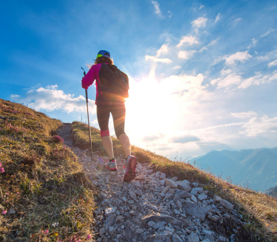 Hiker wearing a pink jacket and blue cap with a backpack climbing a rocky mountain trail at sunrise.