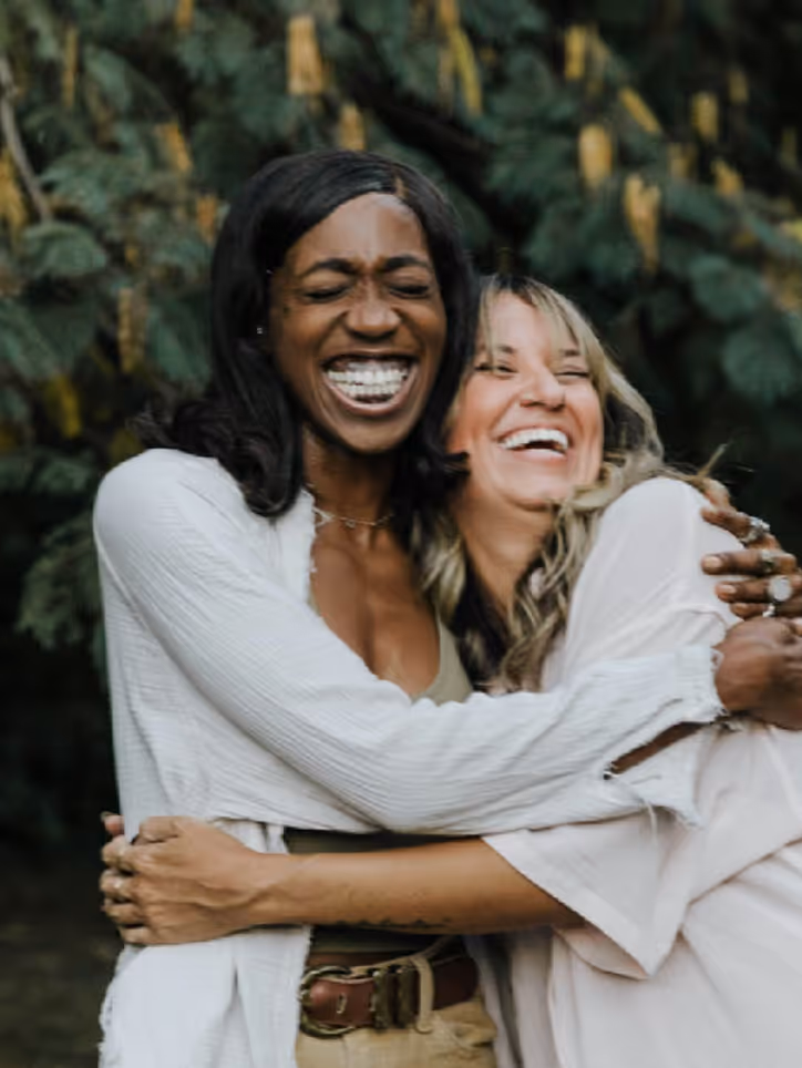 Two women smiling and hugging each other outside with greenery in the background.