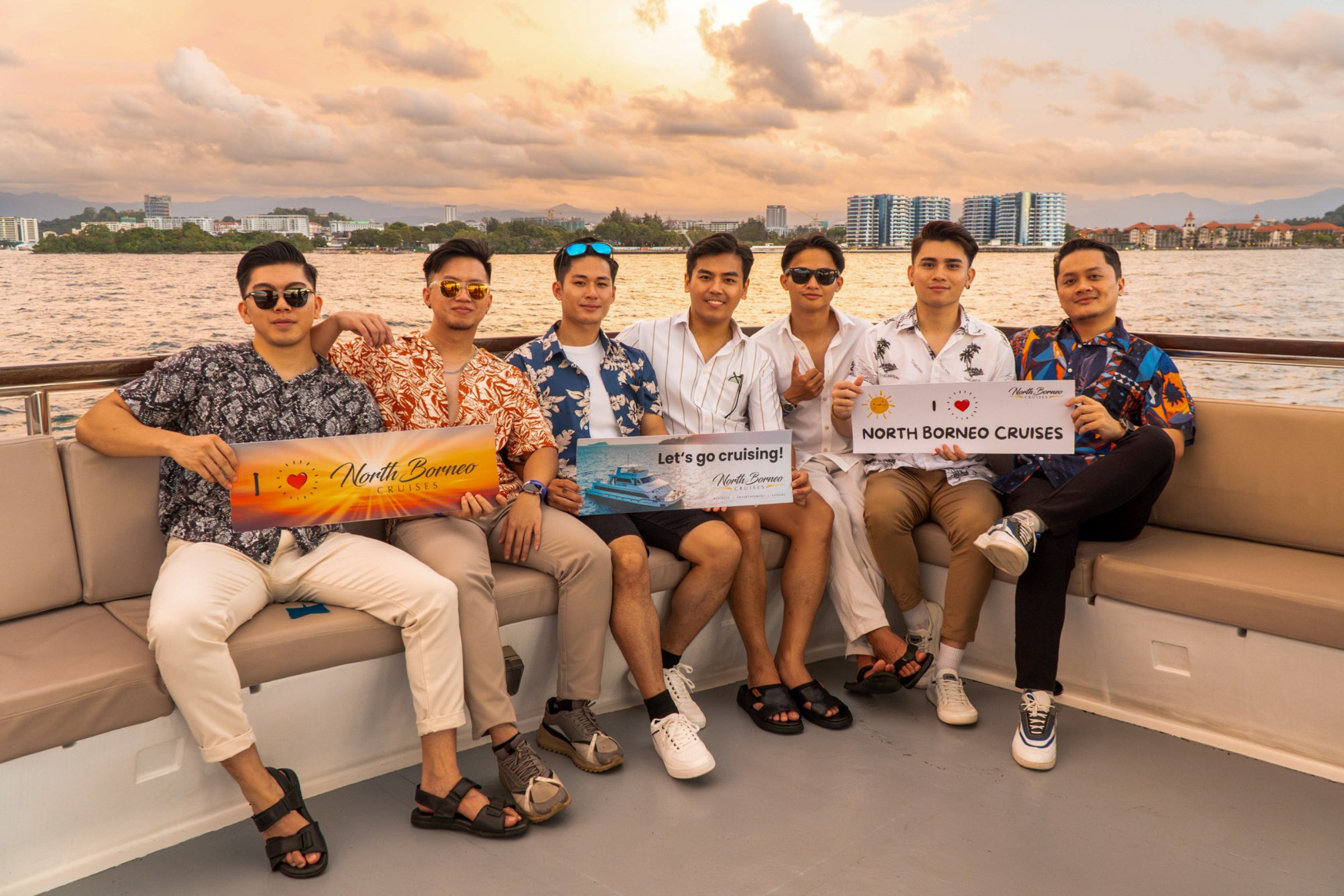 Guests sitting holding signs promoting North Borneo Cruises with sunset background.