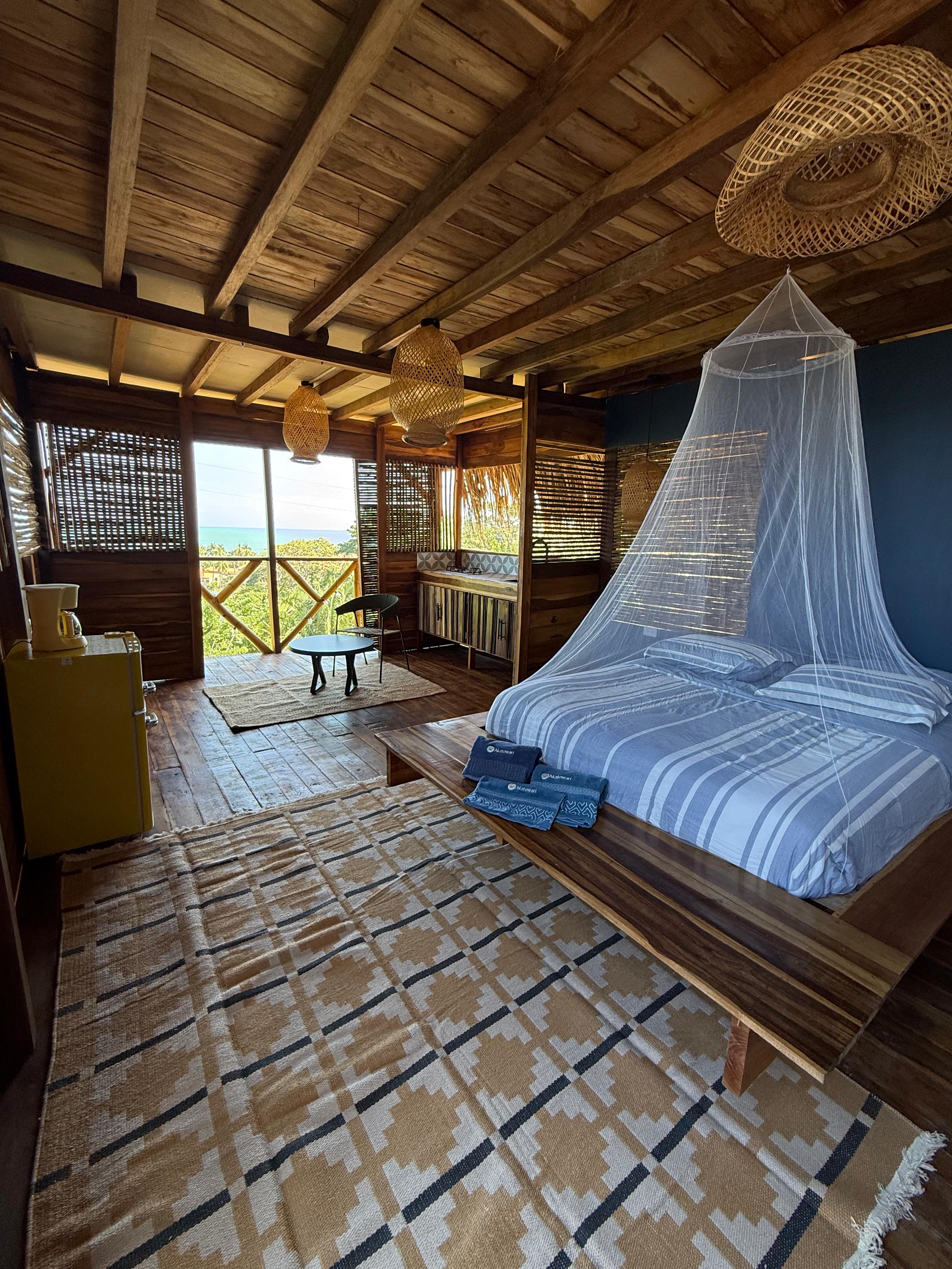 Habitación con cama doble con dos almohadas y mosquitero blanco, decoración rústica de madera, alfombra beige con patrón geométrico, lámparas de mimbre y vista al mar desde un balcón con sofá y mesa pequeña. Akatowari hotel santa marta.