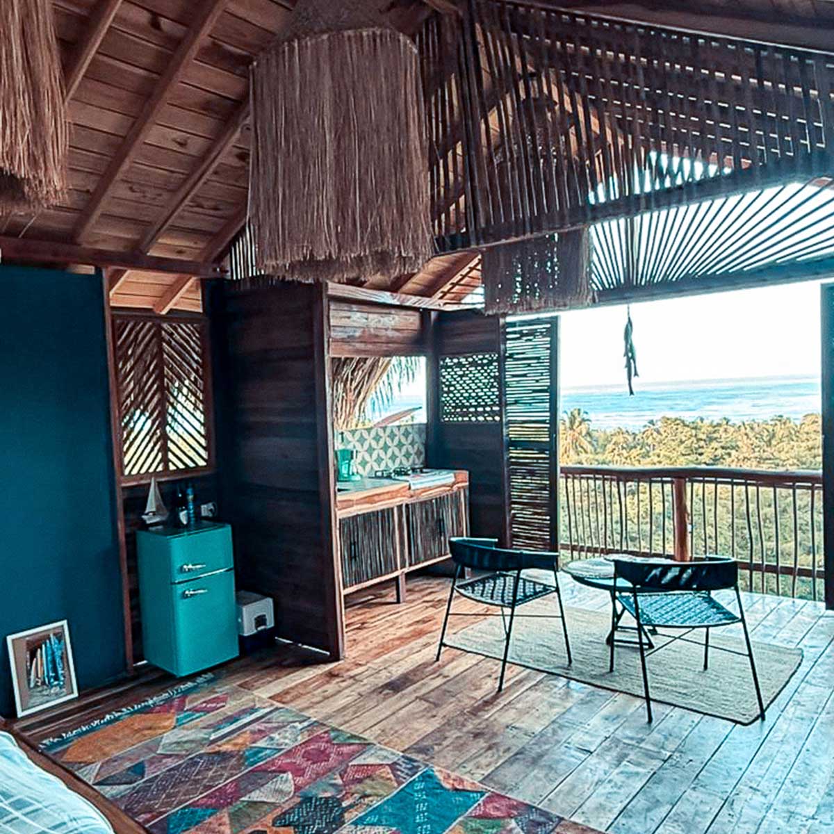 Interior de cabaña con techo de madera, dos sillas negras y mesa frente a balcón con vista a árboles y mar. Akatowari hotel santa marta.