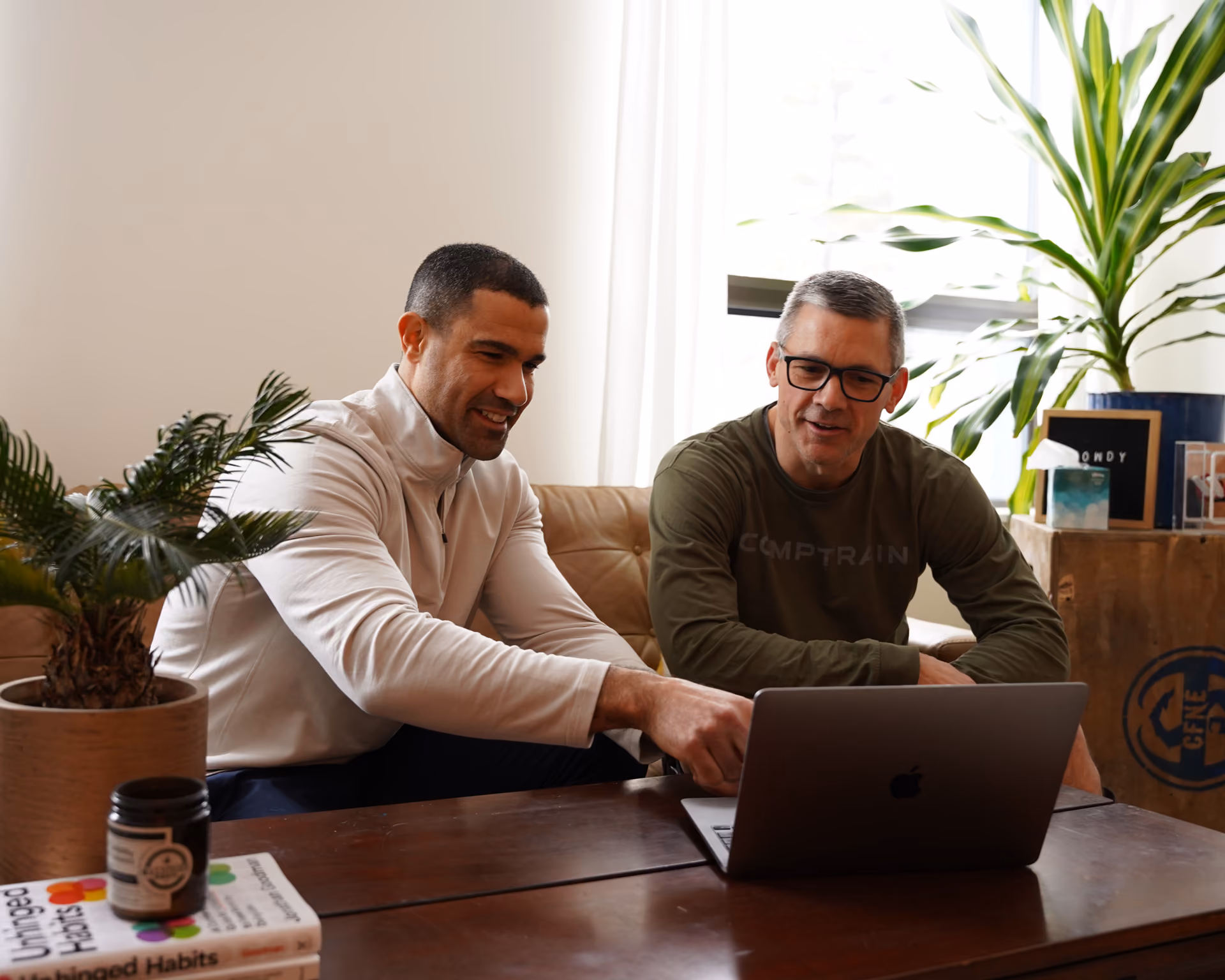 Two men sitting at a table engaged with a laptop in a warmly lit room with plants.