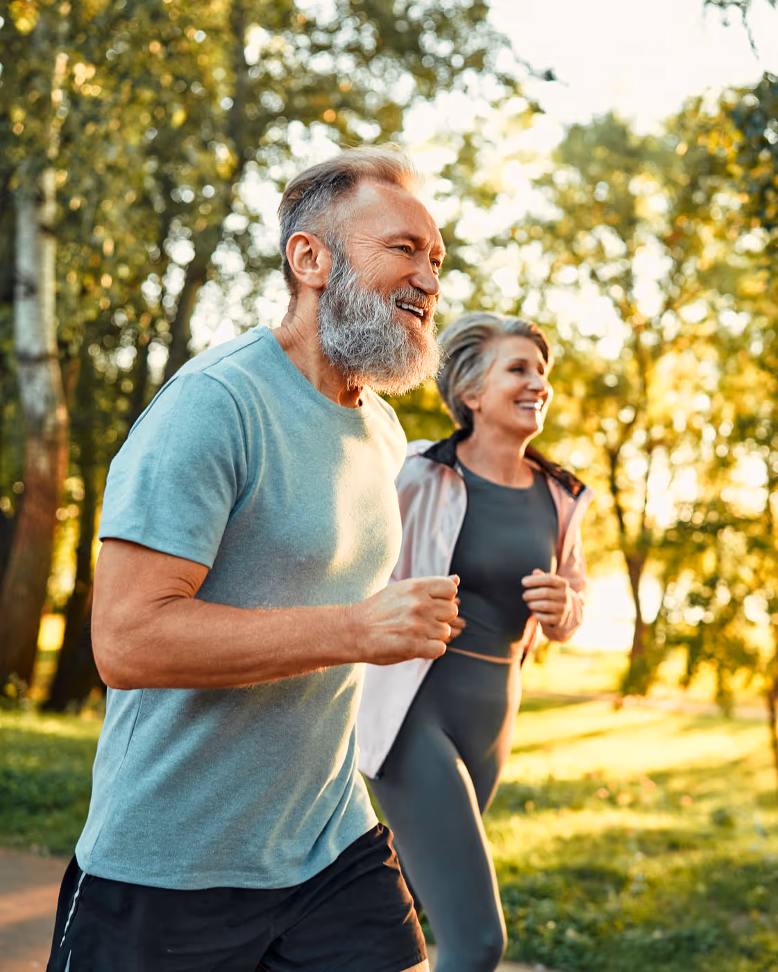 Cardio training outdoors. Side view of grey bearded man and old woman running along summer park with sunlight on background. 