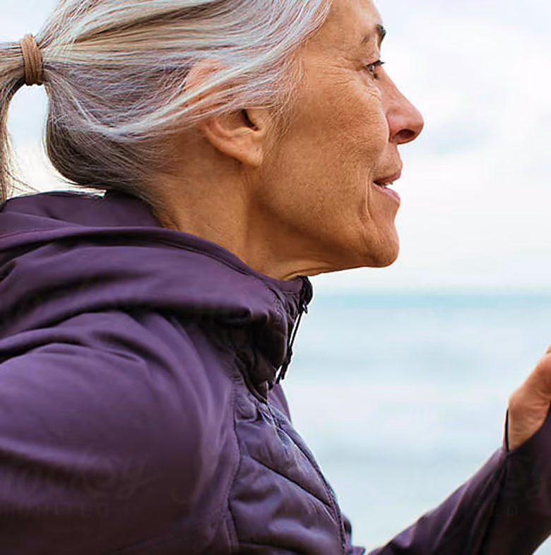Profile of an older woman with gray hair in a ponytail, wearing a purple jacket, outdoors near water.