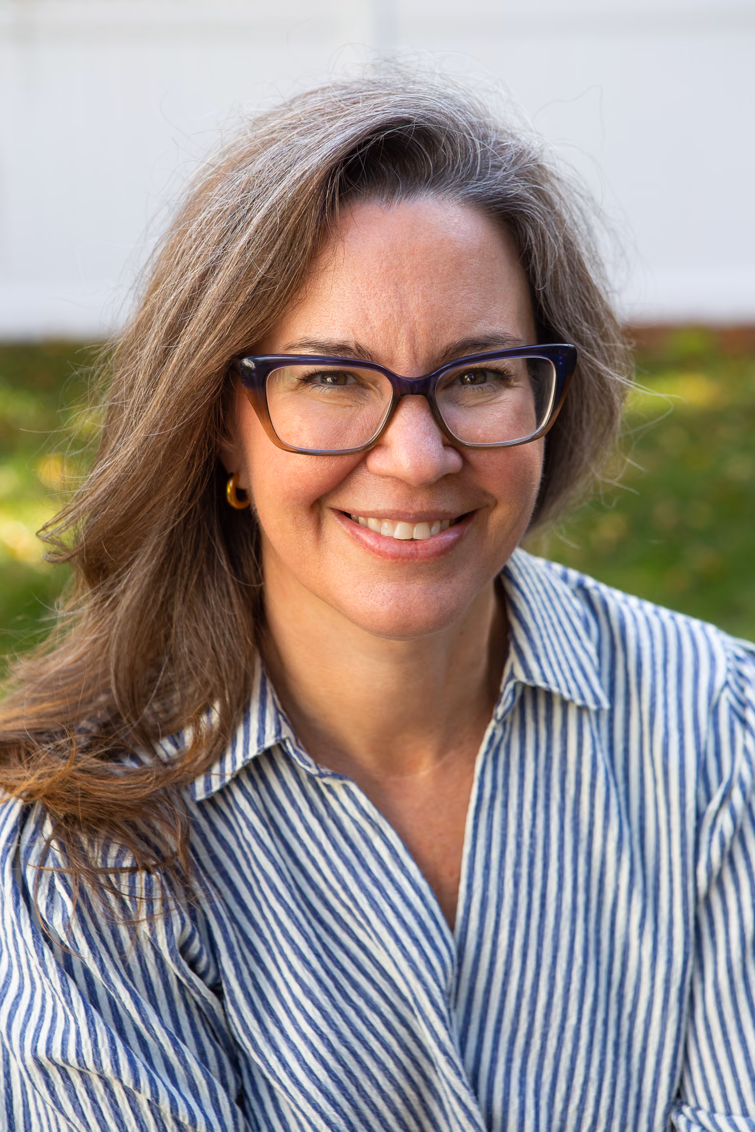 Smiling woman with glasses in a navy blue blouse standing indoors by large green plants.