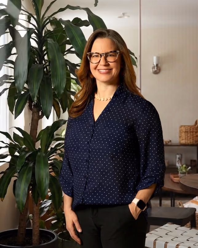 Smiling woman with glasses in a navy blue blouse standing indoors by large green plants.