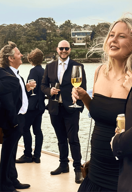 Group of people dressed in formal attire enjoying drinks outdoors near water with greenery and buildings in the background.