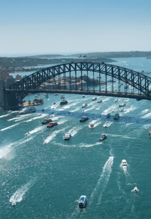 Aerial view of Sydney Harbour Bridge with numerous boats and ferries cruising under it on a clear day.