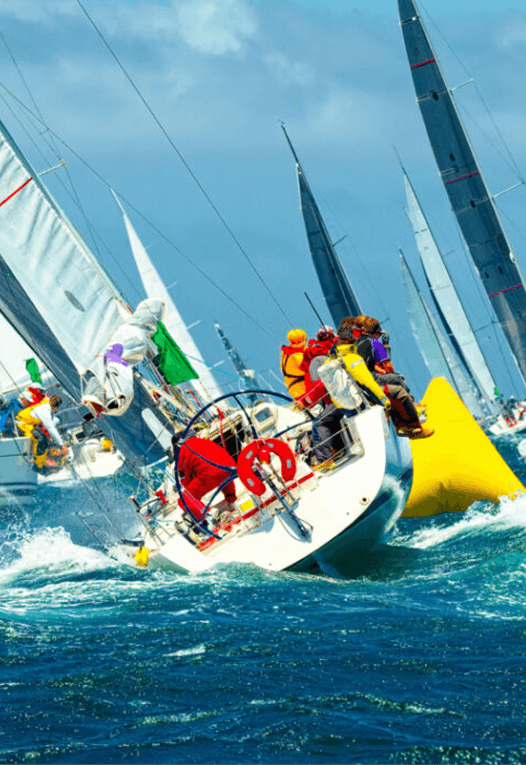 Sailboats with crews in colorful gear racing near a large yellow buoy on a choppy blue sea under a partly cloudy sky.