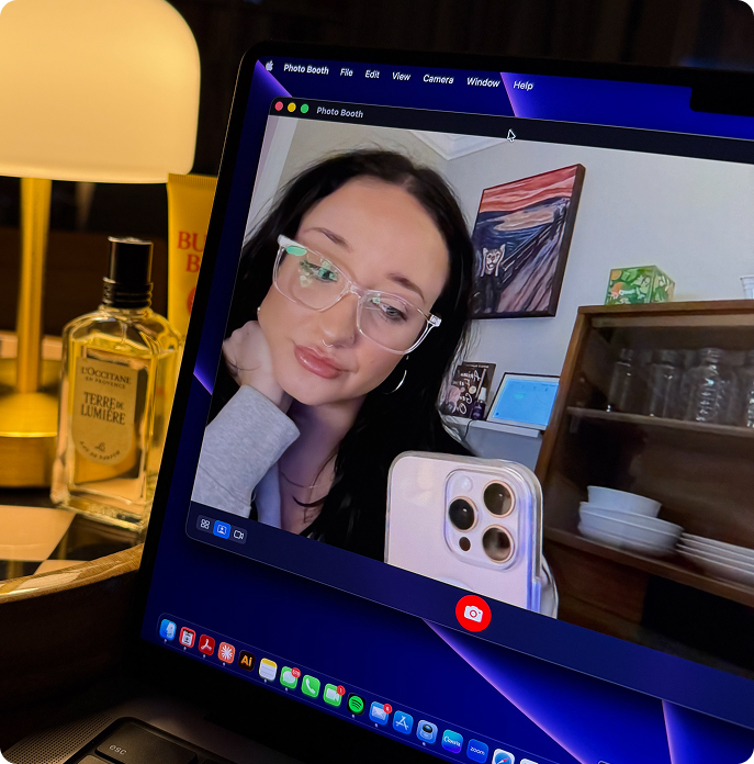 A woman with dark hair and glasses taking a selfie using Photo Booth on a laptop, with a lamp and perfume bottle in the background.