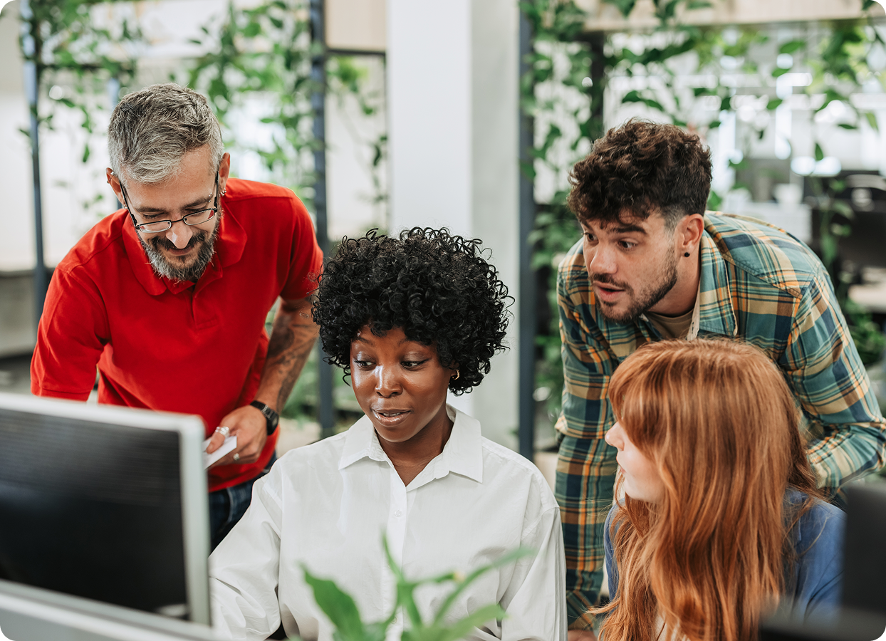 Four diverse coworkers gathered around a computer in an office with green plants, collaborating and discussing.