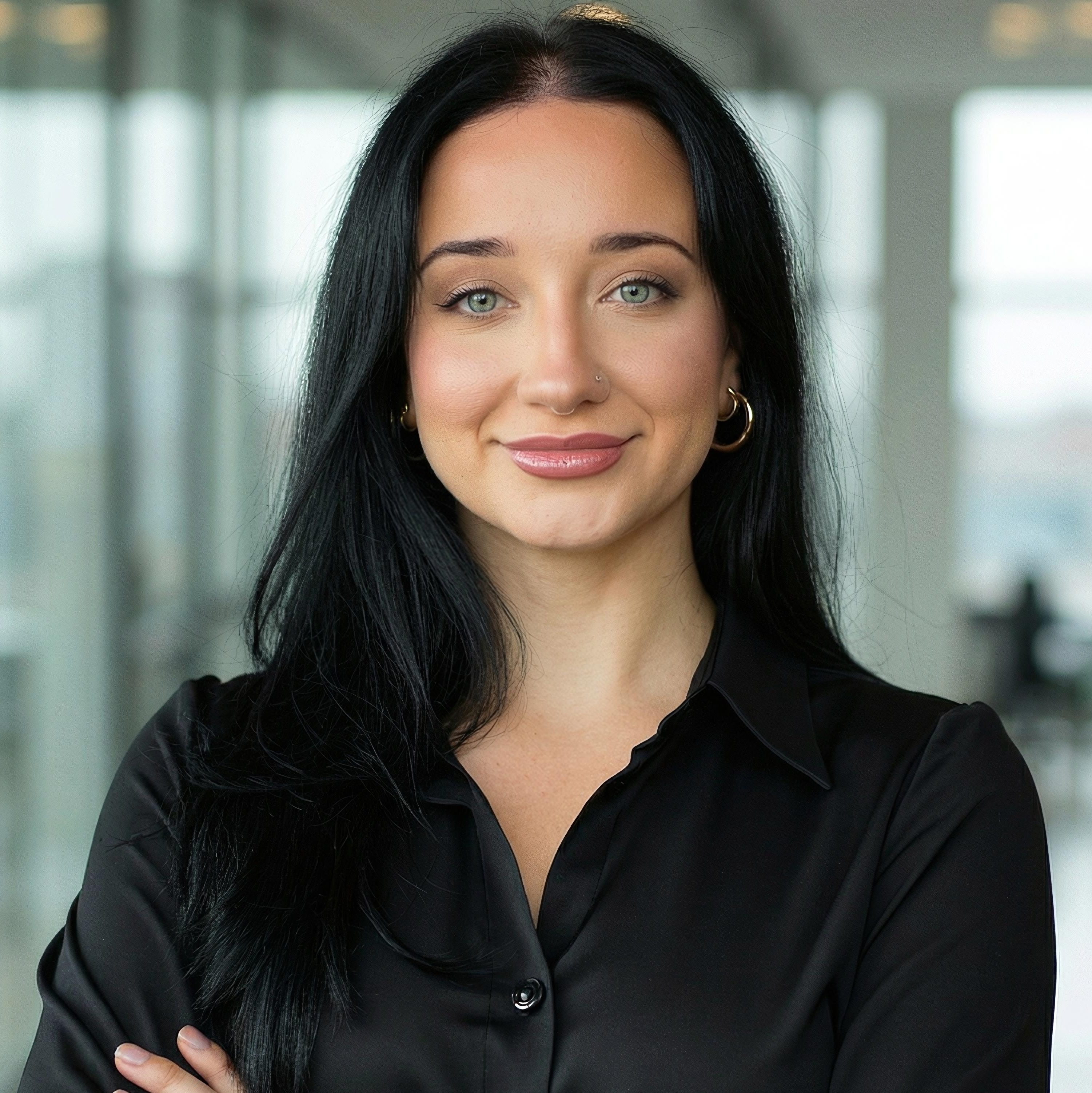 Smiling woman with long black hair, wearing a black button-up shirt and gold hoop earrings, standing in a bright office.