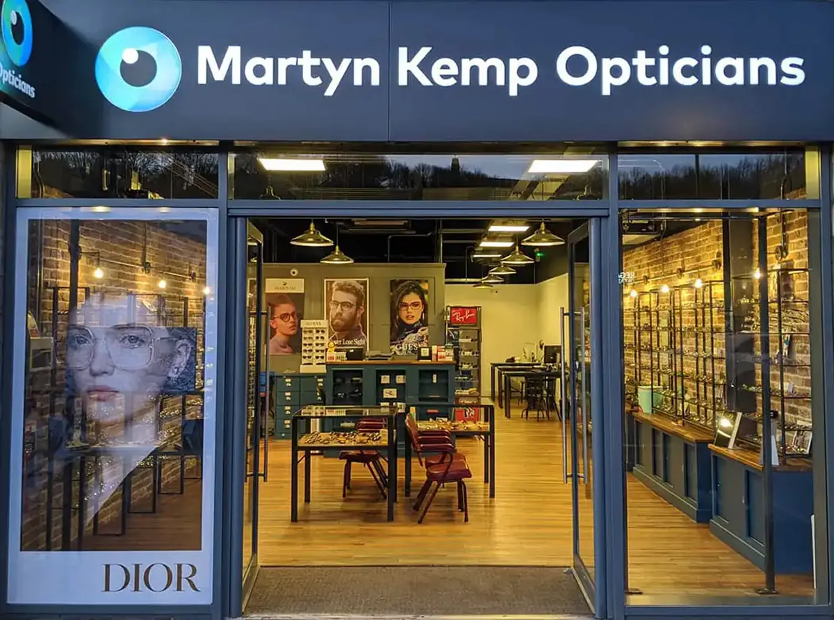 Front view of Martyn Kemp Opticians store with open glass doors, indoor display tables, posters, and racks of eyeglass frames.