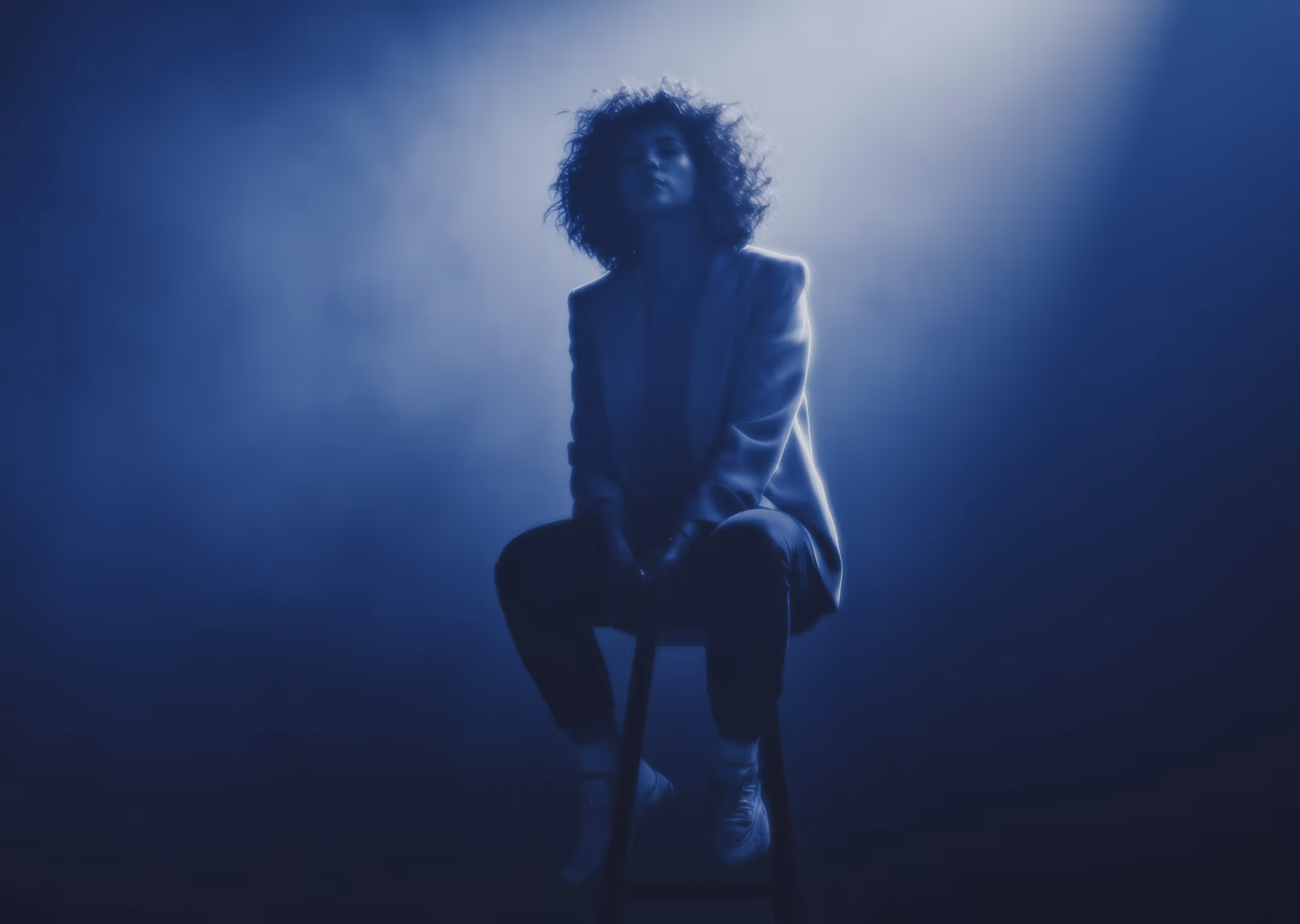 Person with curly hair sitting on a stool, illuminated by dramatic blue backlighting.