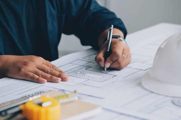 Person drawing architectural plans on blueprints with a pen, with a white construction helmet and measuring tape on the table.