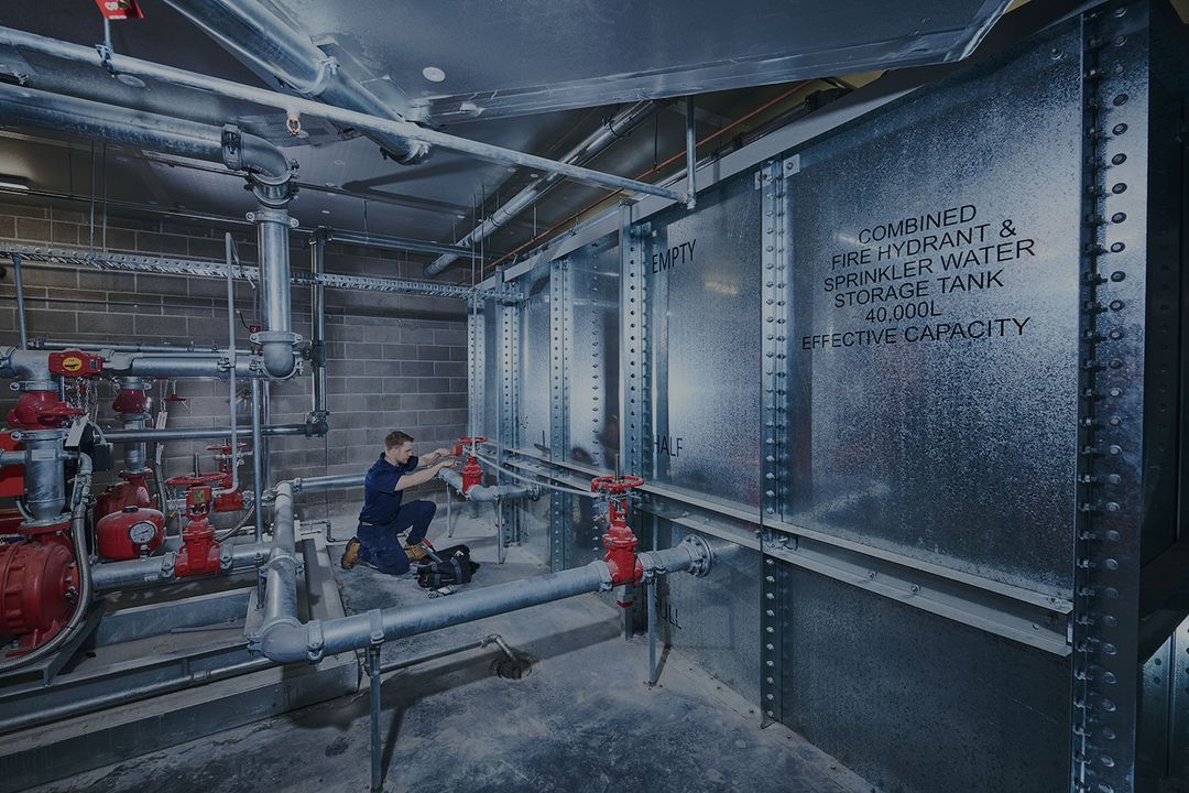 Technician servicing red valves connected to a large metal fire hydrant and sprinkler water storage tank with 40,000L capacity inside an industrial facility.