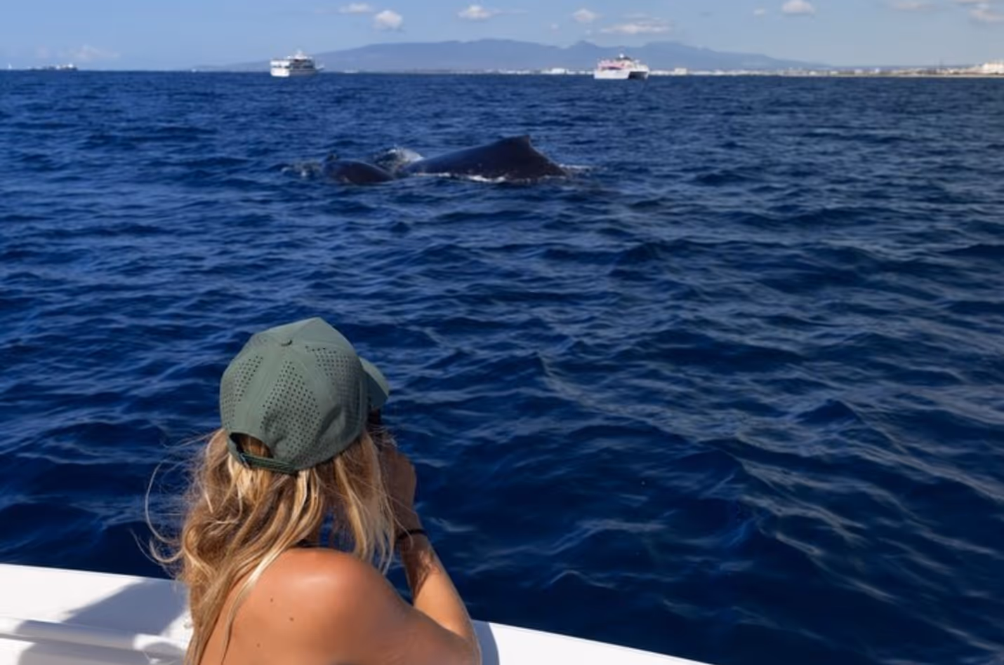A whale can be seen breaching the surface, not too far from the bow of a boat, while a woman looks on with her camera. 