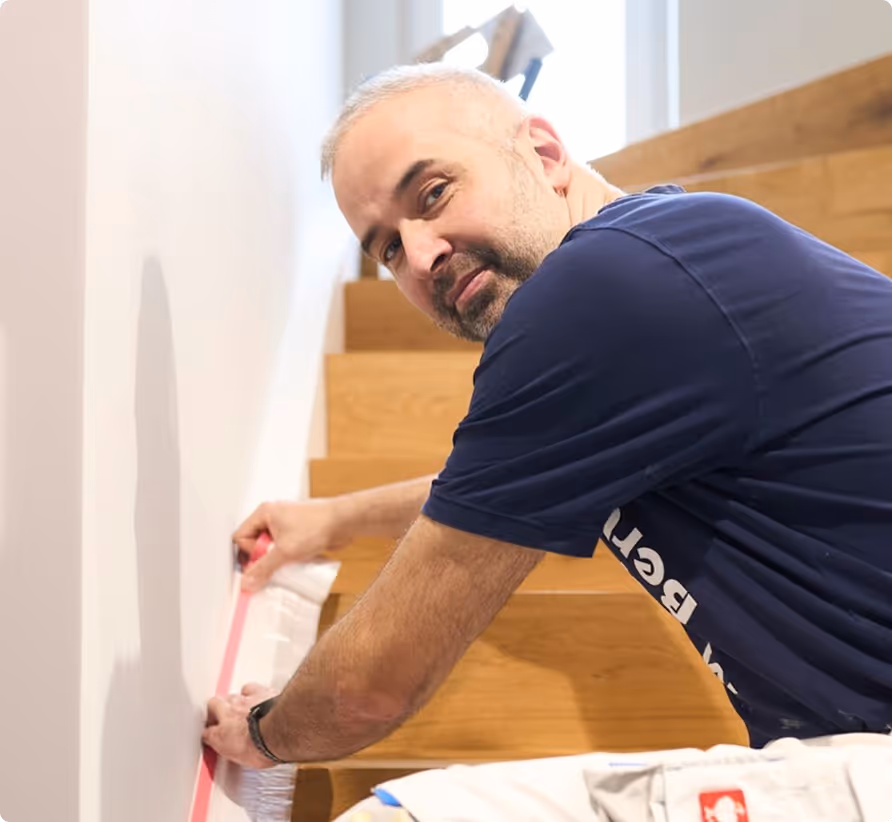 Ein Mann misst mit einem Maßband eine Wand neben einer Holztreppe.