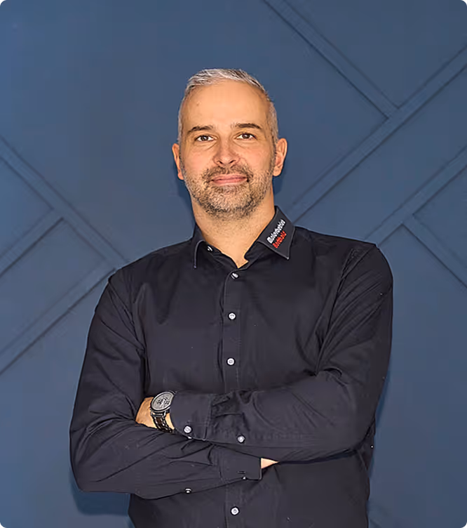 Mann mit kurzen grauen Haaren und Bart, trägt ein schwarzes Hemd mit verschränkten Armen vor blauem Hintergrund.