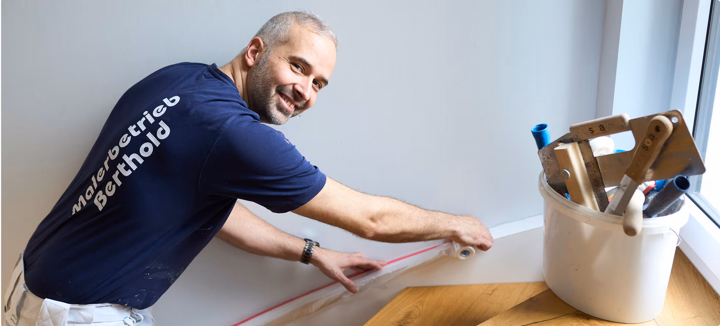 Mann in blauem T-Shirt mit der Aufschrift 'Malereibetrieb Berthold' klebt Schutzfolie an eine Wand, daneben ein Eimer mit Malerwerkzeugen.