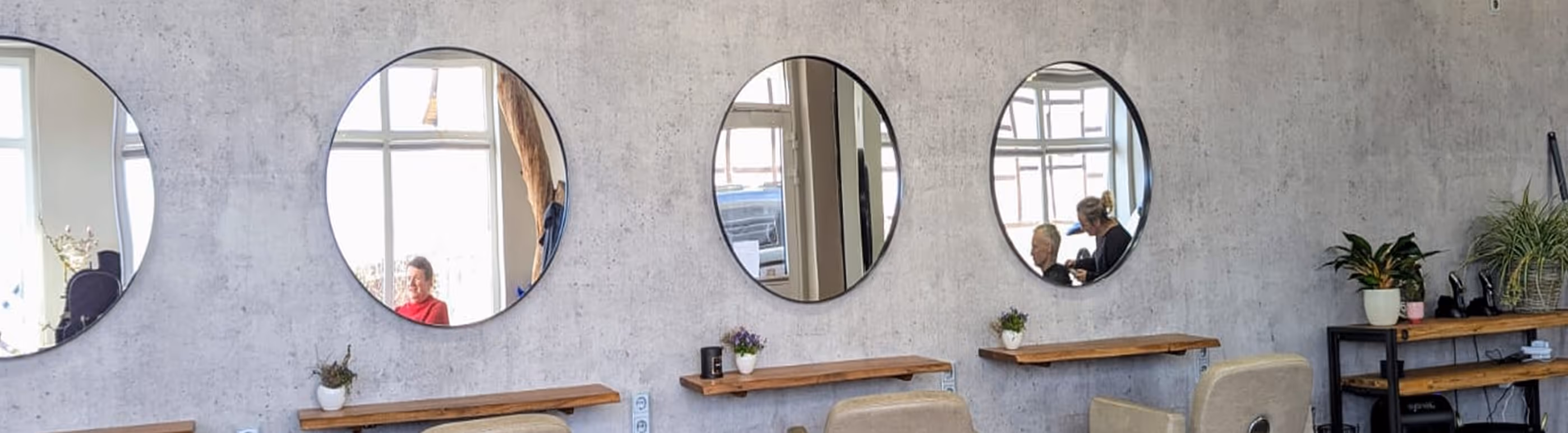 Friseursalon mit vier runden Spiegeln an einer grauen Wand, Holzablagen darunter und Haarstylistin, die einem Kunden die Haare schneidet.