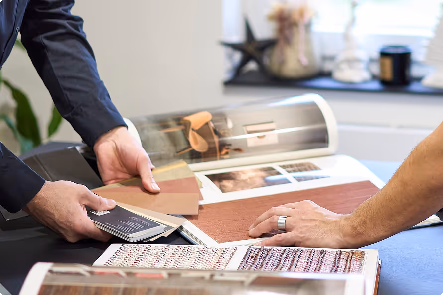 Zwei Personen vergleichen Farbmuster und Holzmuster auf einem Tisch mit geöffneten Musterbüchern.