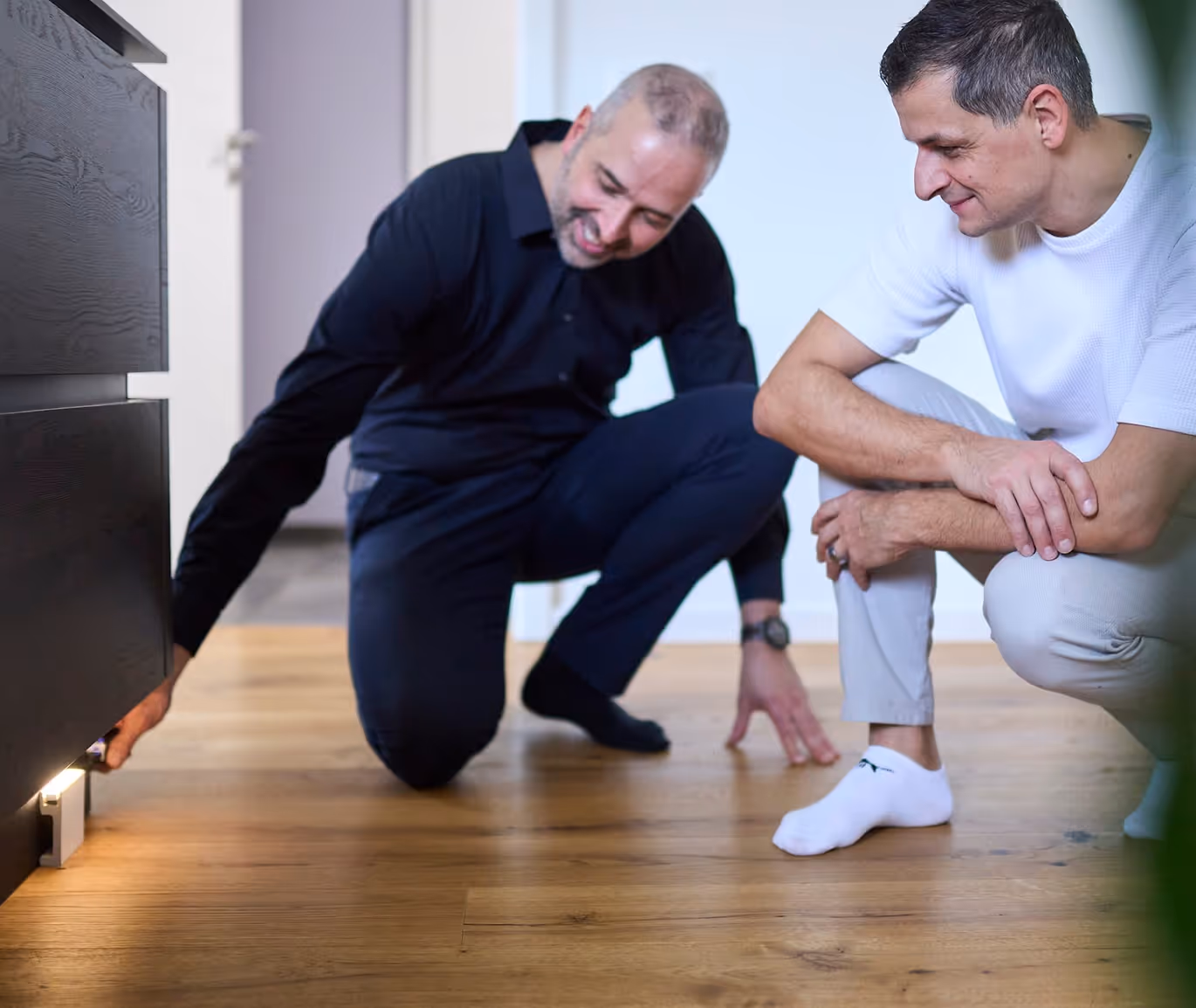 Zwei Männer knien auf einem Holzboden, einer prüft eine unter einem Möbelstück angebrachte Lichtquelle.
