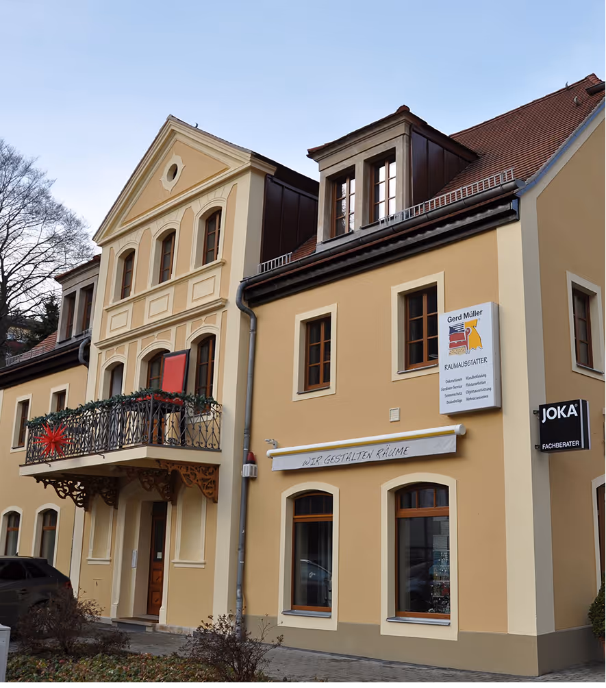 Traditionelles gelbes Gebäude mit verziertem Balkon und zwei Schildern an der Fassade, eines mit der Aufschrift 'Gerd Müller Raumausstatter'.
