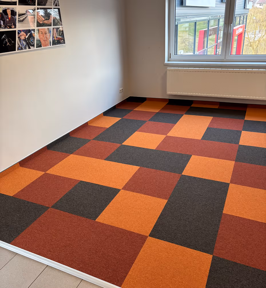 Räumliches Büro mit braun-orange-grauen quadratischen Teppichfliesen, weißer Wand, Fenster und Heizung.