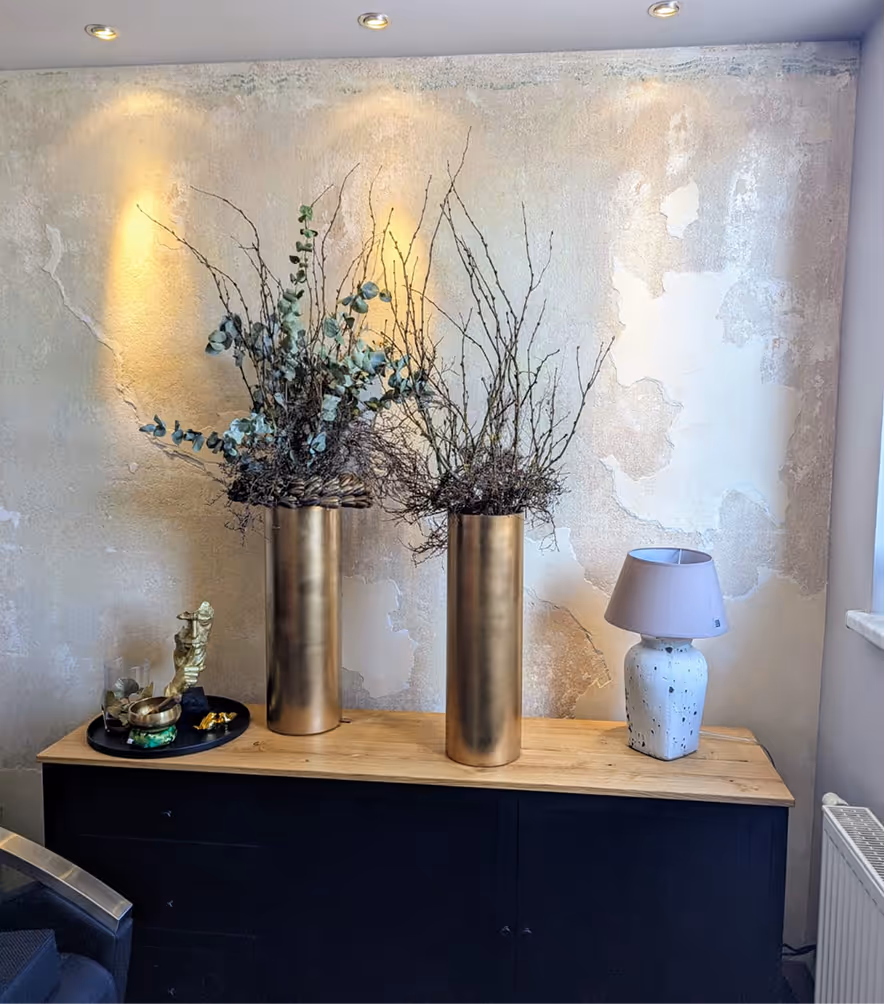 Zwei goldene Vasen mit getrockneten Zweigen und Blättern auf einem Holzschrank neben einer weißen Keramiklampe mit grauem Lampenschirm vor einer strukturierten Wand.