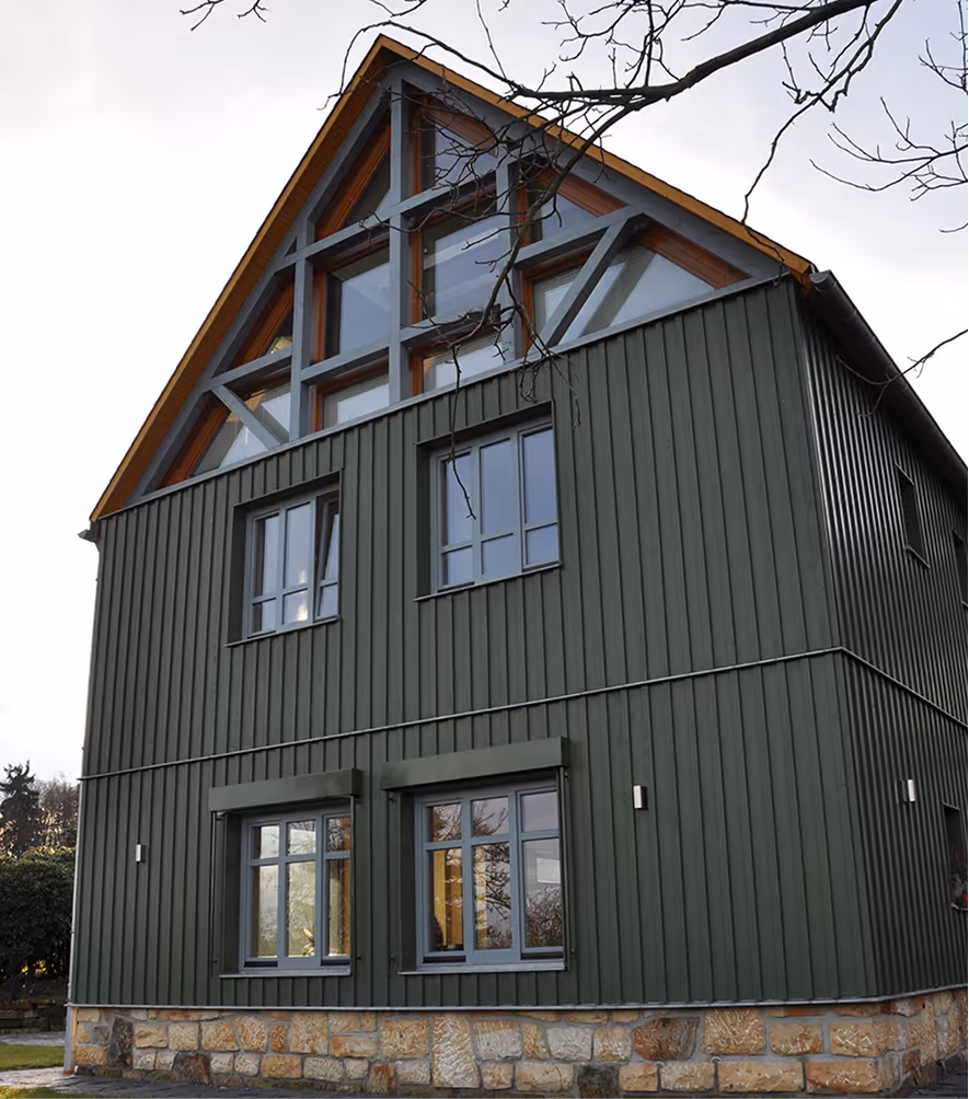 Modernes zweistöckiges Haus mit dunkelgrüner vertikaler Holzverkleidung und großen Fenstern unter einem dreieckigen Dach mit Holz- und Metallrahmen.