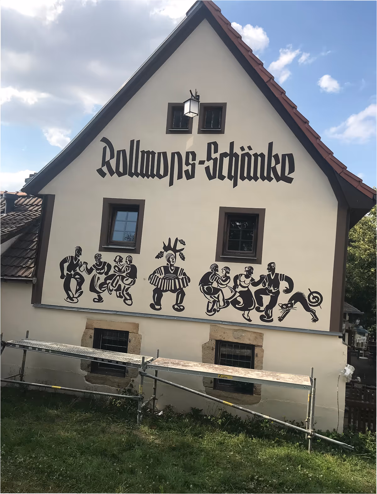 Hausfassade mit der Aufschrift ‚Rollmops-Schänke‘ und schwarzen Zeichnungen von tanzenden Menschen und einer Katze.