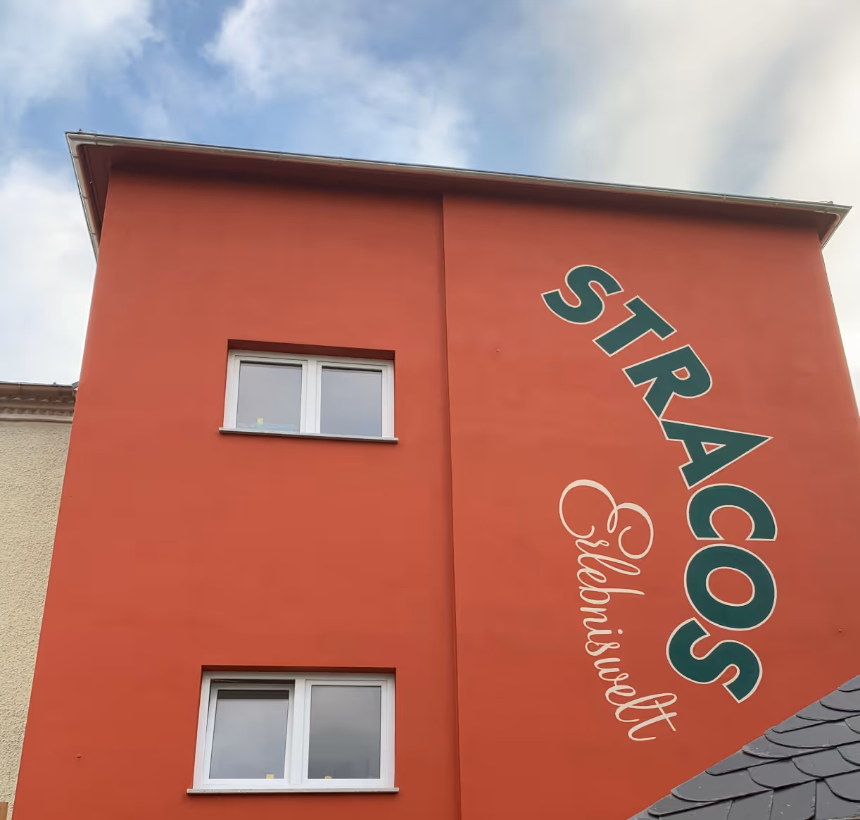 Rotes Gebäude mit zwei weißen Fenstern und dem diagonalen Schriftzug ‚STRACOS Eiseniswelt‘ an der Fassade.