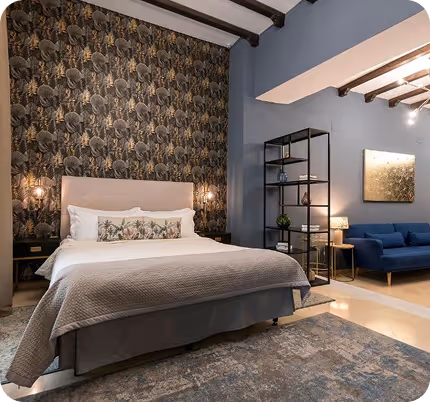 Modern bedroom with a large bed featuring patterned pillows, floral wallpaper behind the headboard, a blue sofa, black metal shelf unit, and a large area rug.