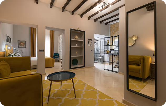 Modern living room with yellow furniture, patterned rug, glass coffee table, and visible bedroom and kitchen areas.