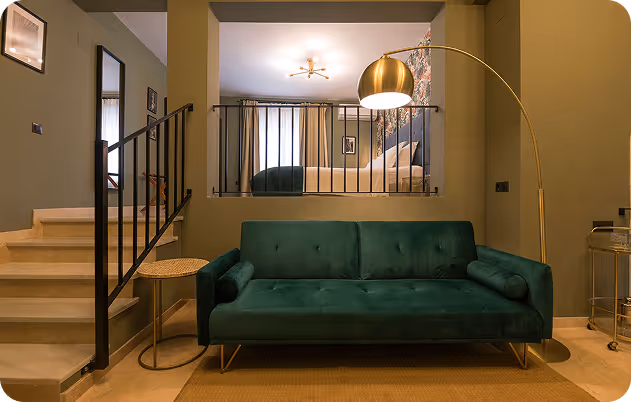Modern living area with a green velvet sofa, gold floor lamp, and a view into a raised bedroom with floral wallpaper.
