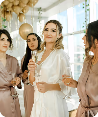 Bride in white robe holding a champagne glass with bridesmaids in matching robes at an indoor celebration.