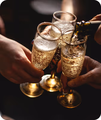 Three hands holding champagne flutes clinking glasses while champagne is being poured into one glass.