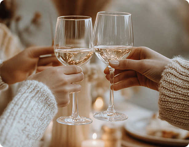 Two people wearing knitted sweaters clinking glasses filled with white wine in a cozy setting.