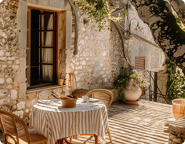 Cozy outdoor patio with a striped tablecloth-covered round table, two wicker chairs, stone wall, and sunlight casting shadows.