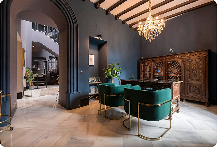 Elegant room with dark blue walls, two green velvet chairs with gold frames, a wooden desk, antique cabinet, crystal chandelier, and arched doorway leading to a staircase.