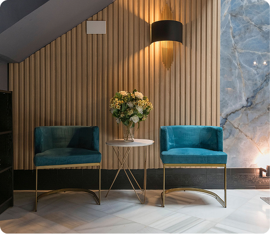 Two teal velvet chairs with gold legs facing each other, separated by a round white table with a vase of flowers, set against a wooden slat wall and marble panel.