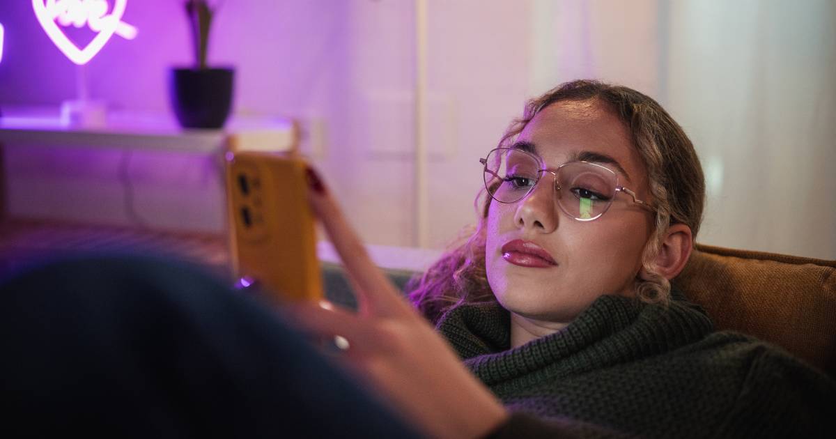 Young woman relaxing on a comfortable sofa, browsing social media on her smartphone in a cozy, neon-lit living room.