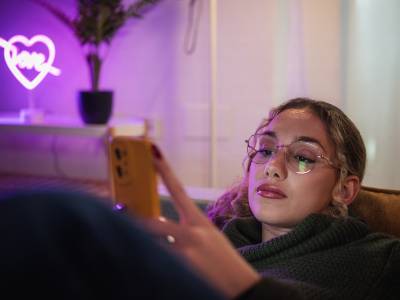 Young woman relaxing on a comfortable sofa, browsing social media on her smartphone in a cozy, neon-lit living room.